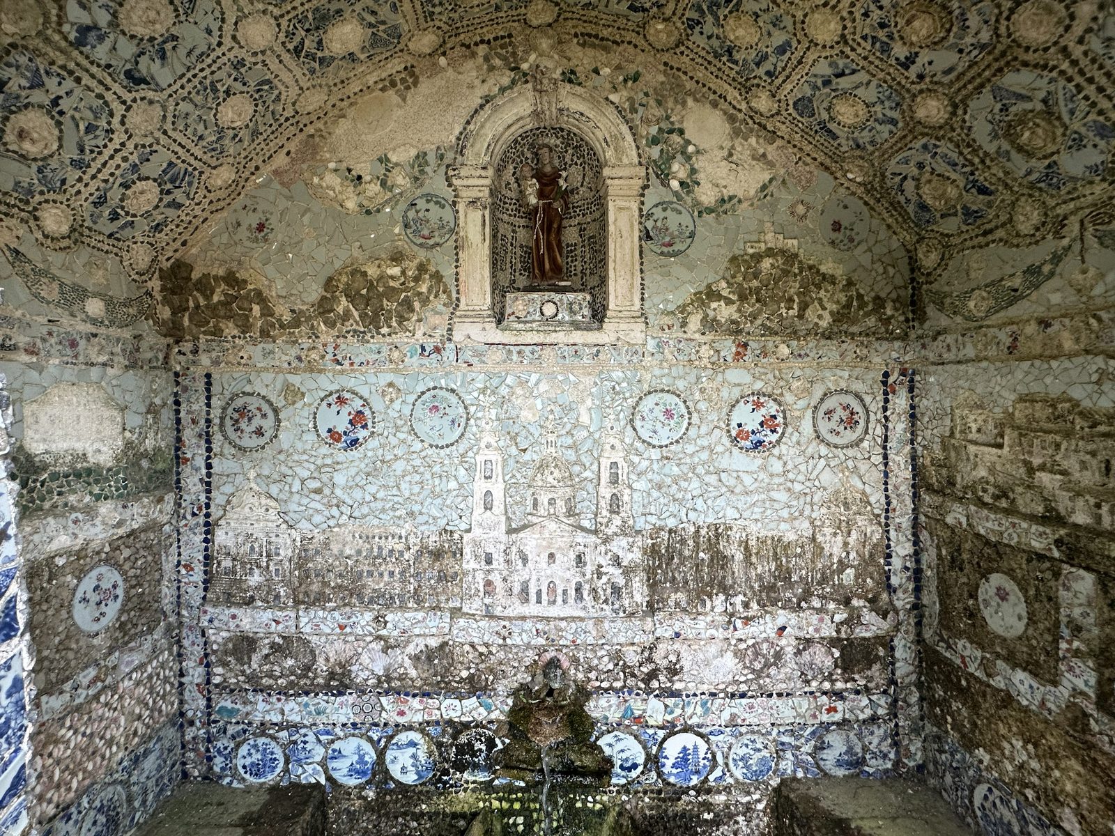 The Mosaic Niche — Shell-work and old tiles in a garden alcove, watched over by a small saint.